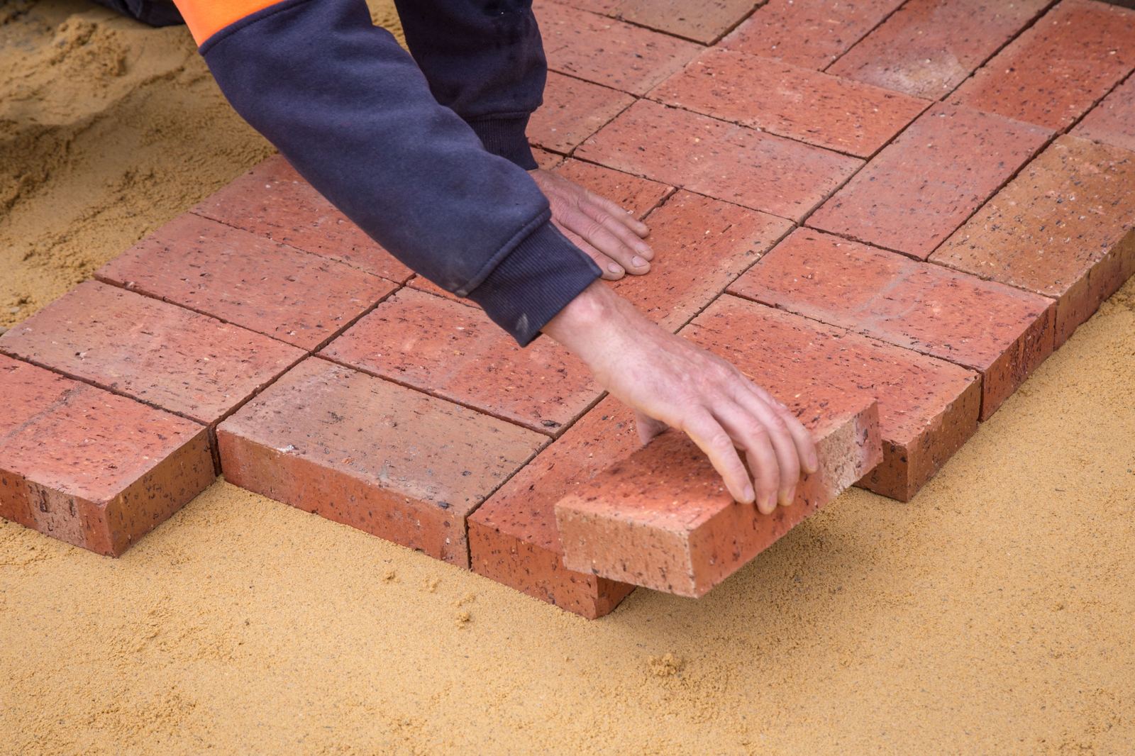 Hands placing red brick pavers into a sand base