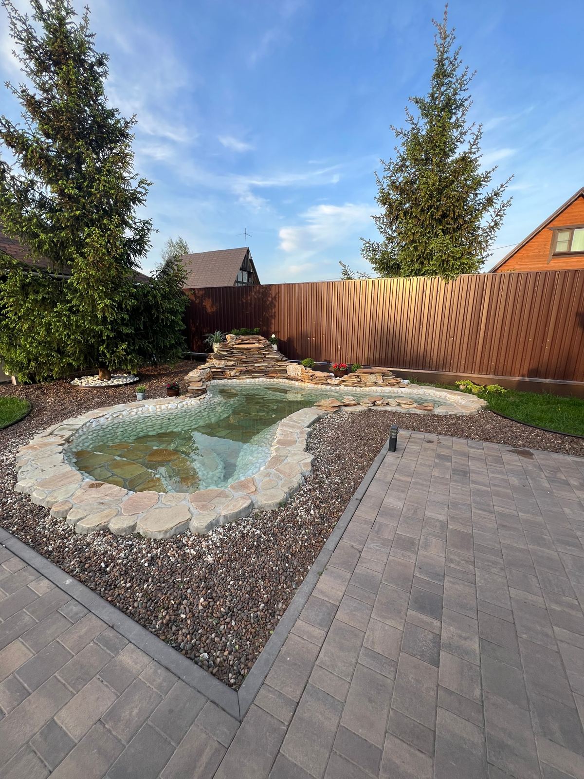 Backyard stone-edged pond with a paver patio and mature pine trees