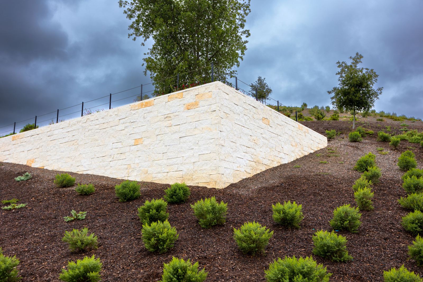 Tall stacked stone retaining wall on a sloped hillside lot under a dramatic sky