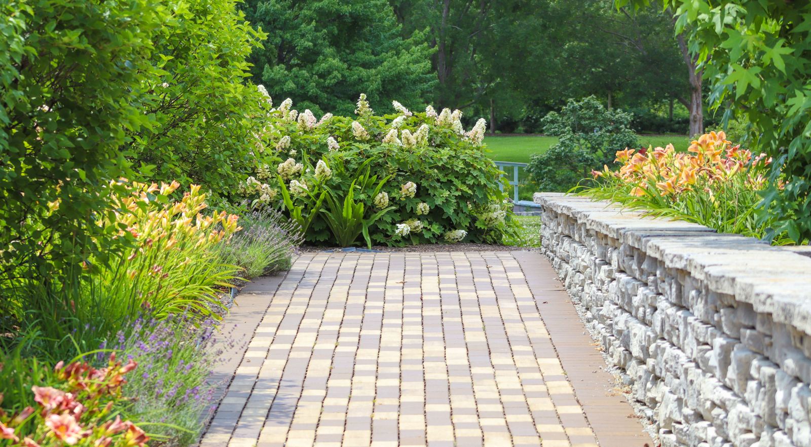 Paver walkway running alongside a stacked stone retaining wall and flower beds